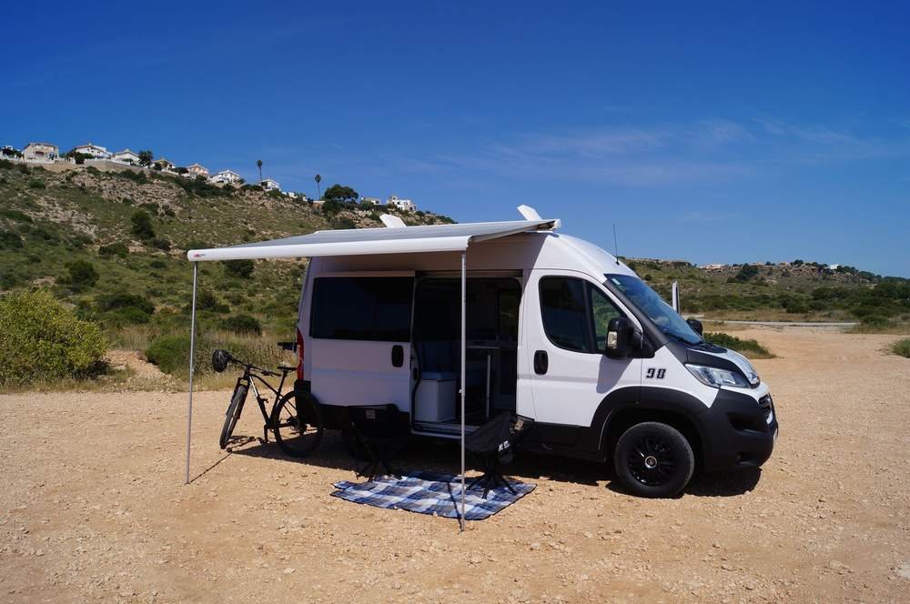 Furgoneta camper de Domo Vans con el toldo desplegado y una bicicleta, frente al monte y el mar en Alicante